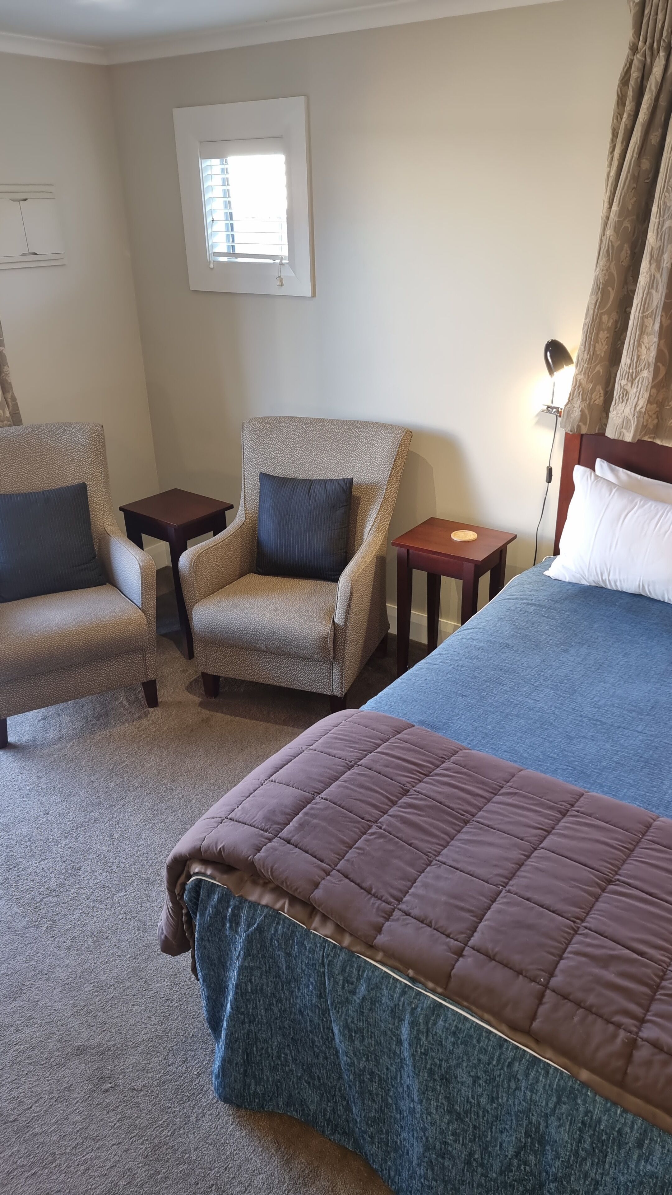 A hotel room with a bed, two armchairs, a small table, a lamp, and a window with blinds.