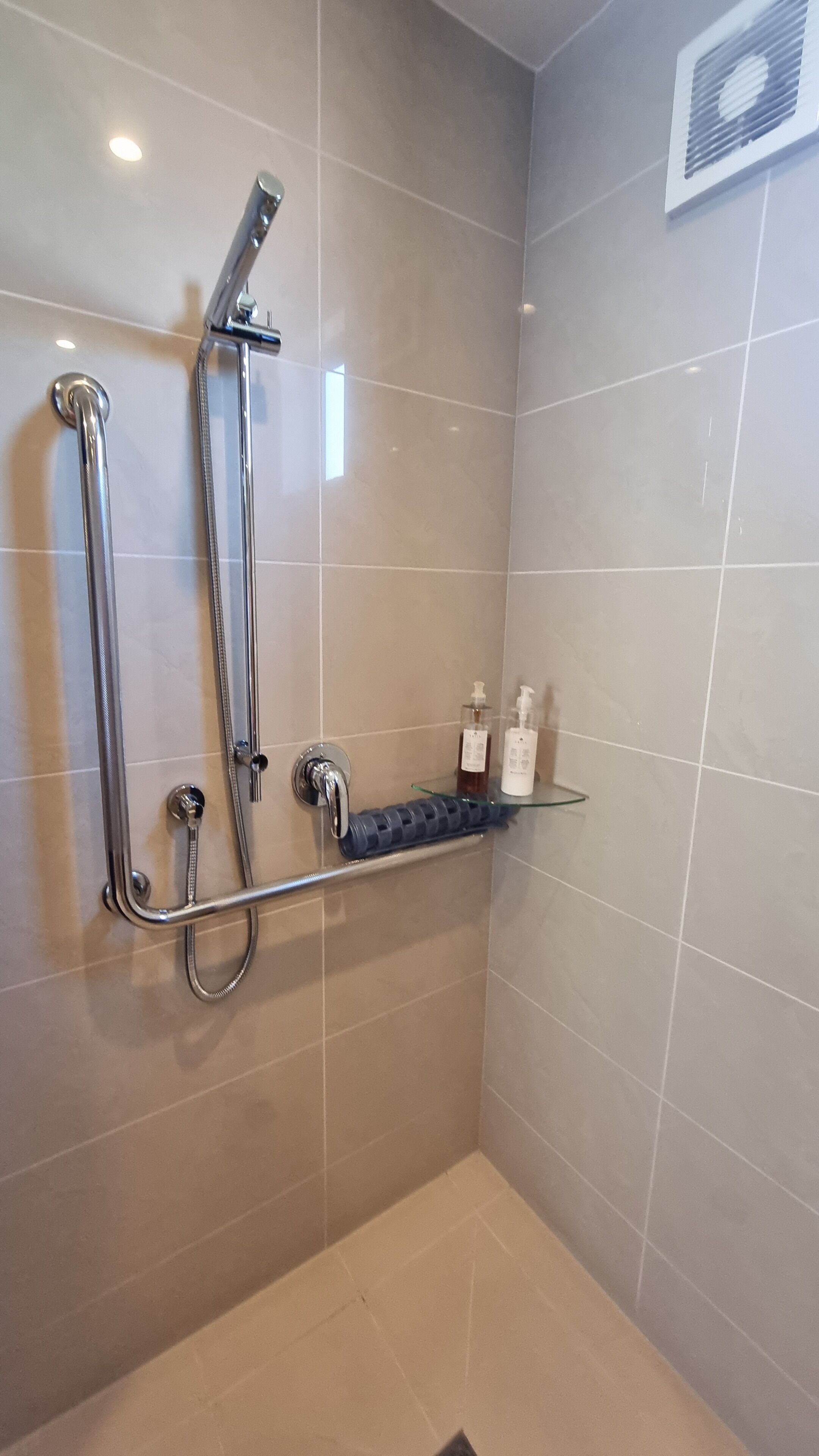 A shower area with a grab bar, a handheld showerhead, and a glass shelf with toiletries.