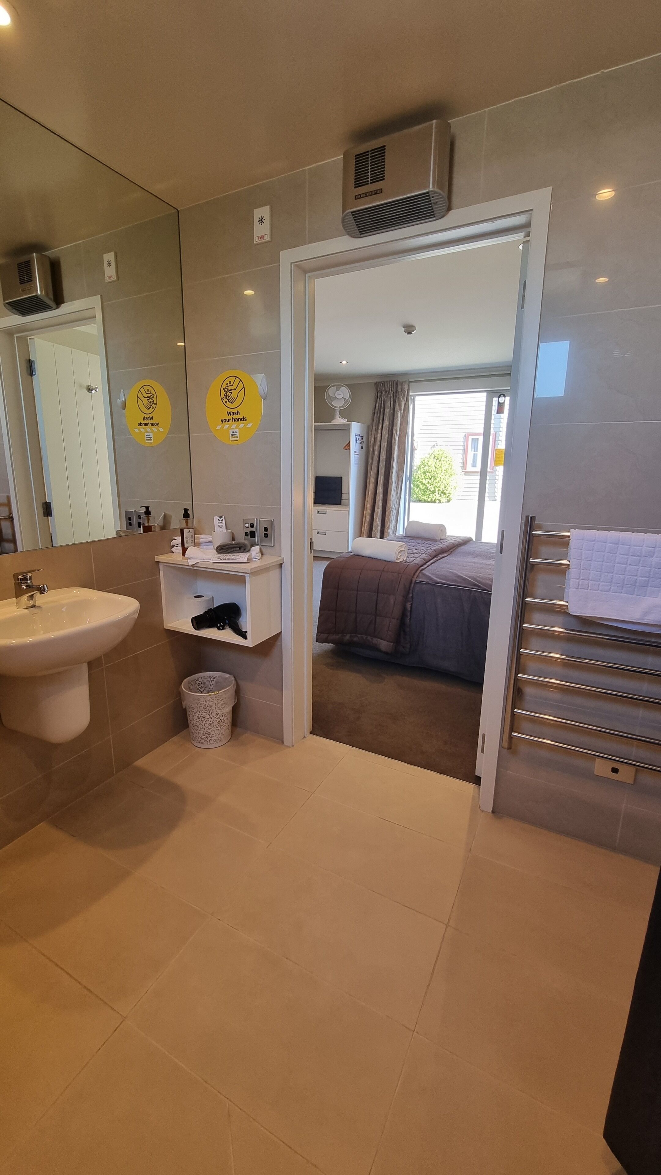 A modern bathroom with a sink, mirror, and toilet. There are hand sanitizer dispensers and a trash bin. The bathroom leads to a bedroom with a bed and a window.