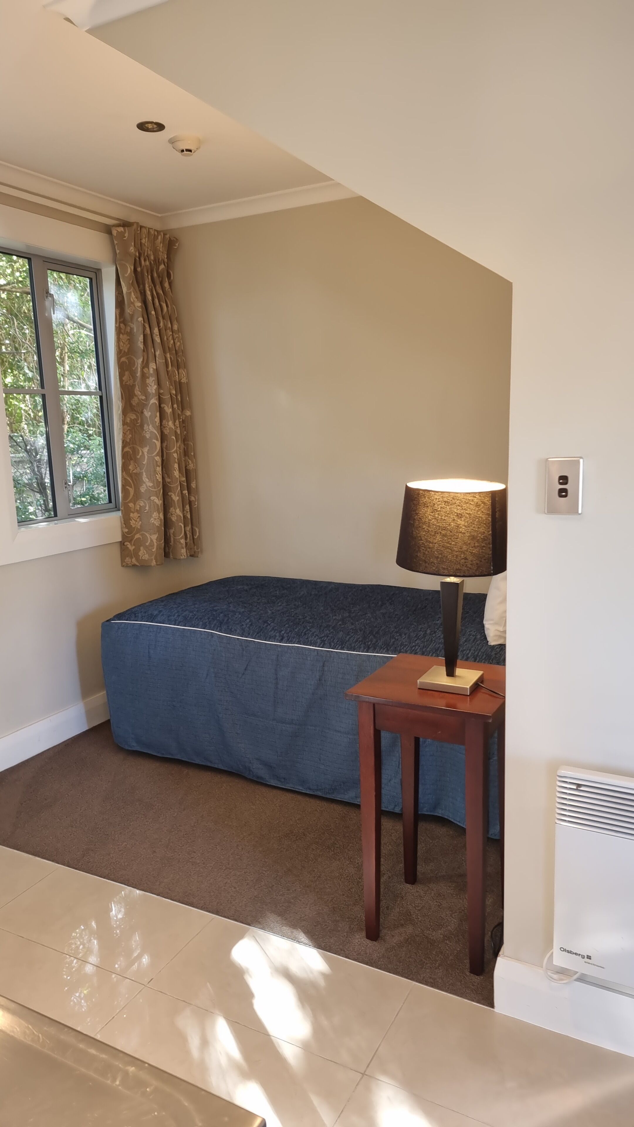 Une chambre à coucher avec un lit, une lampe, un radiateur et une fenêtre avec des rideaux.