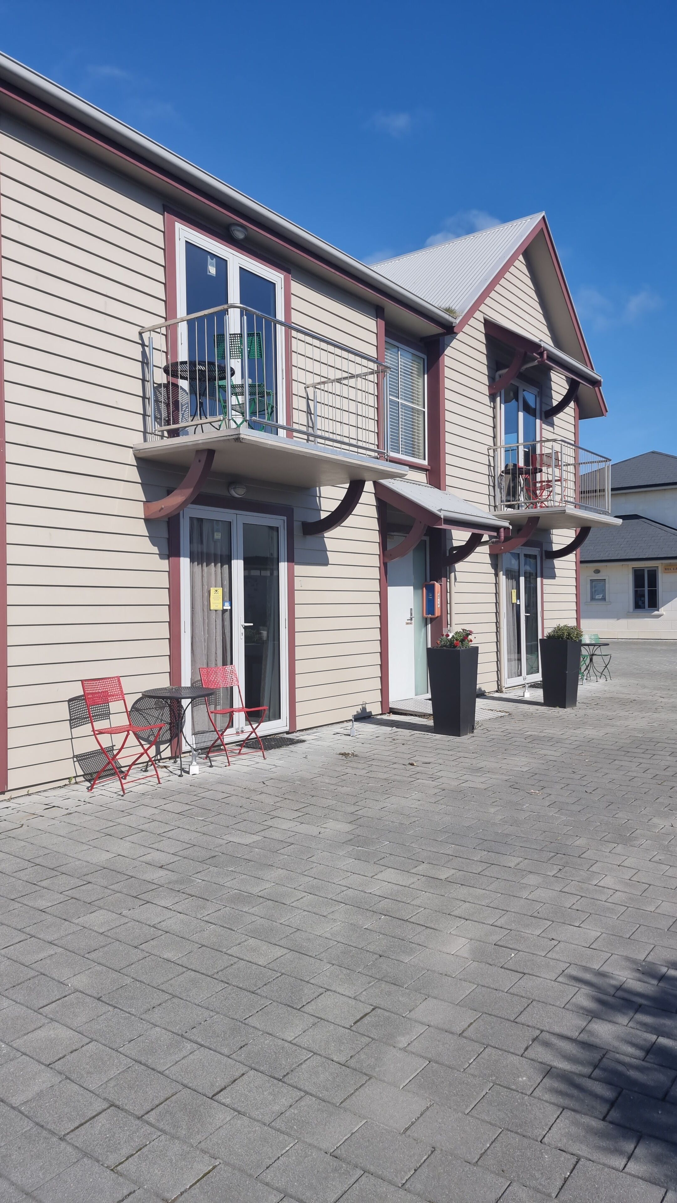 A building with a flat roof, a balcony with metal railings, and a covered entrance area with two red chairs and a table.