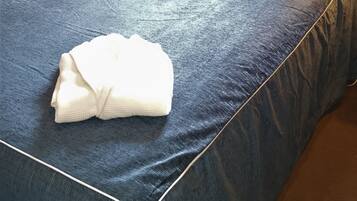 Una cama bien tendida con una colcha azul, una toalla blanca doblada y dos almohadas, una con estampado de hojas.