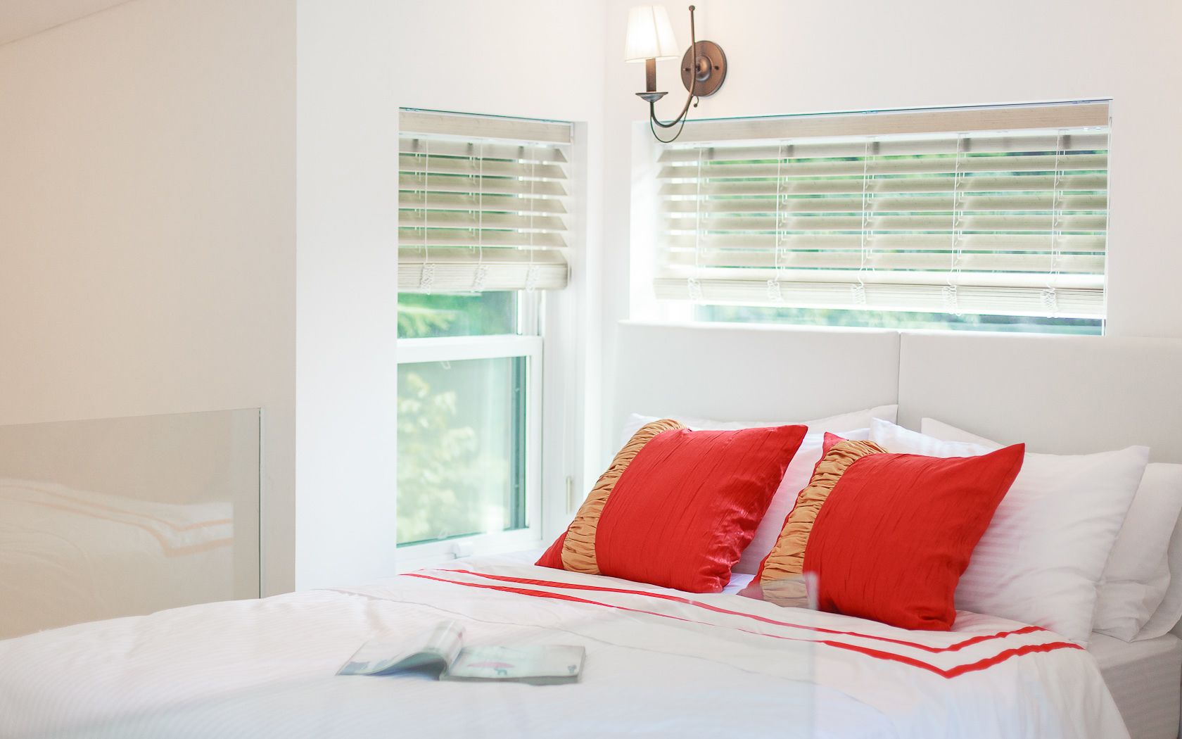 A bedroom with a bed, two red pillows, a white headboard, and a window with blinds.