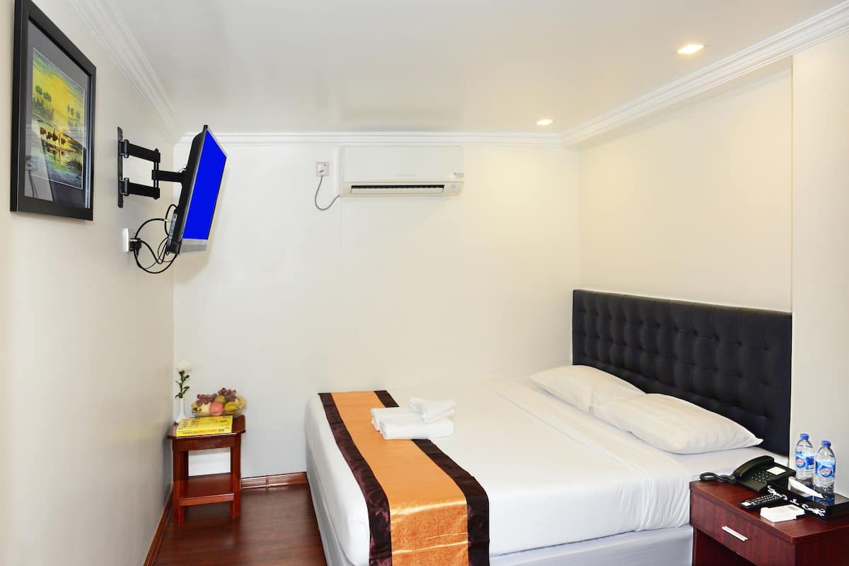 A hotel room with a bed, a wall-mounted TV, an air conditioner, a bedside table with a vase and fruit, and a nightstand with a phone and water bottles.