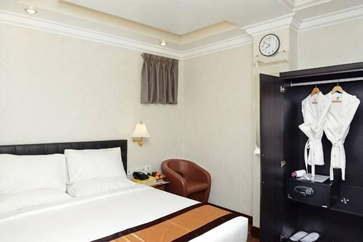 A hotel room with a bed, a chair, a wardrobe, and a clock on the wall.