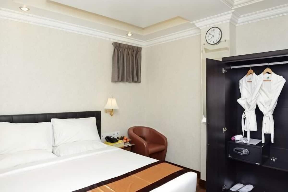 Hotellihuone, jossa on sänky, tuoli, vaatekaappi ja seinällä oleva kello.