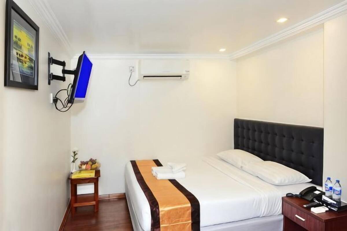 A hotel room with a bed, a bedside table with a phone and water bottles, a TV mounted on the wall, and an air conditioning unit.