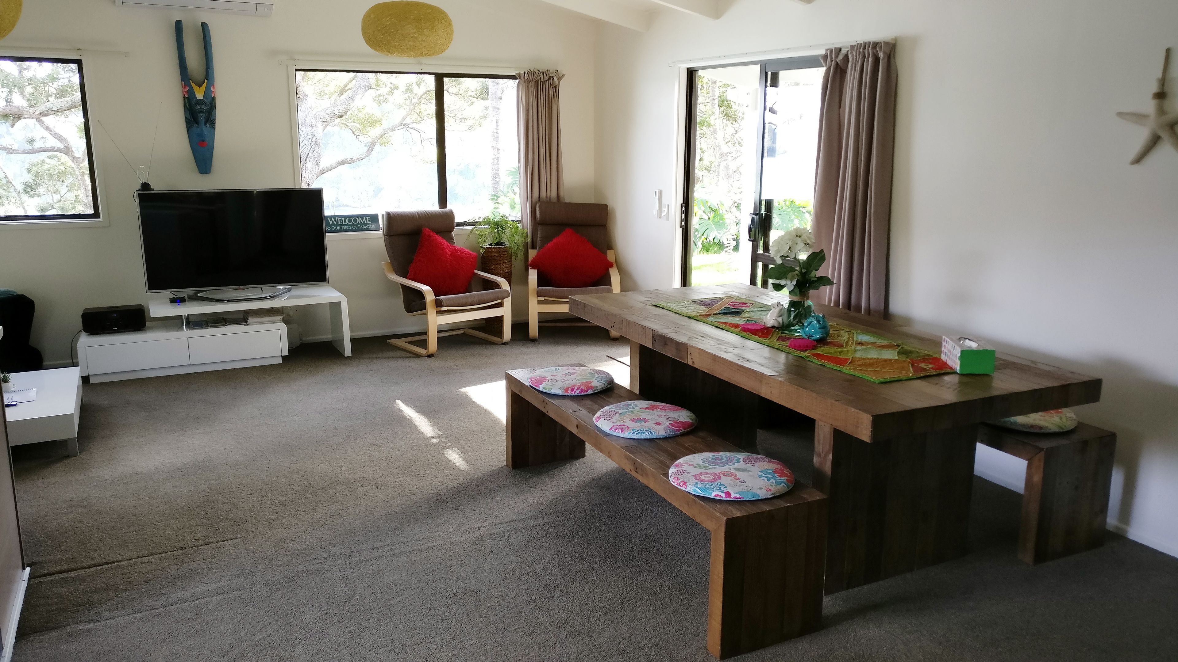 A modern living room with a flat-screen TV, a wooden dining table with benches, and a comfortable seating area with red cushions.