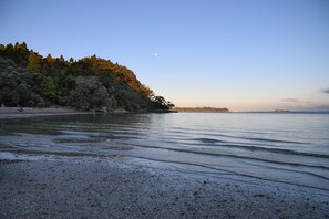 Una playa con aguas tranquilas, cielo despejado y una colina boscosa al fondo.
