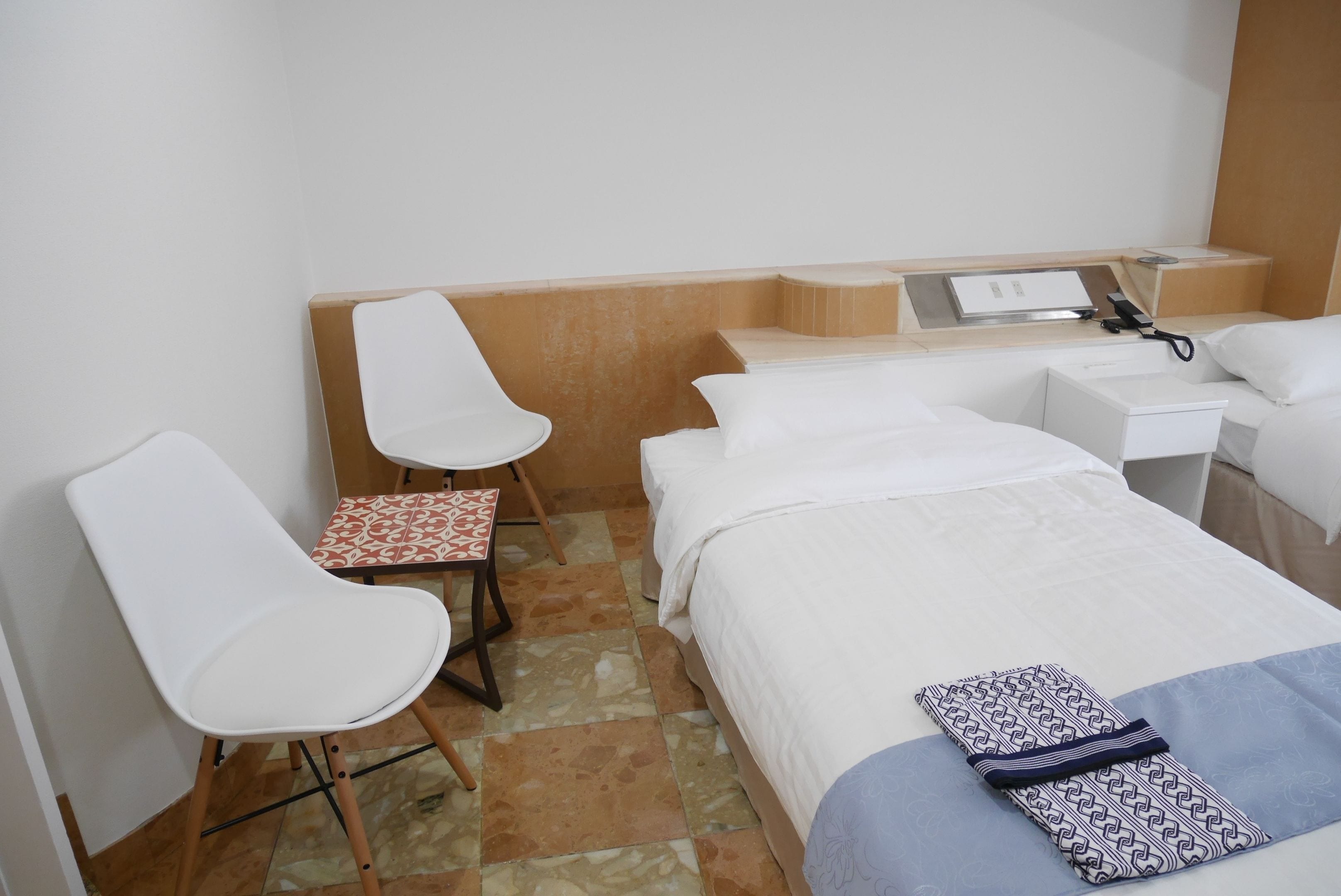Una habitación de hotel pequeña con una cama, una silla, una mesita y una mesita de noche con lámpara.