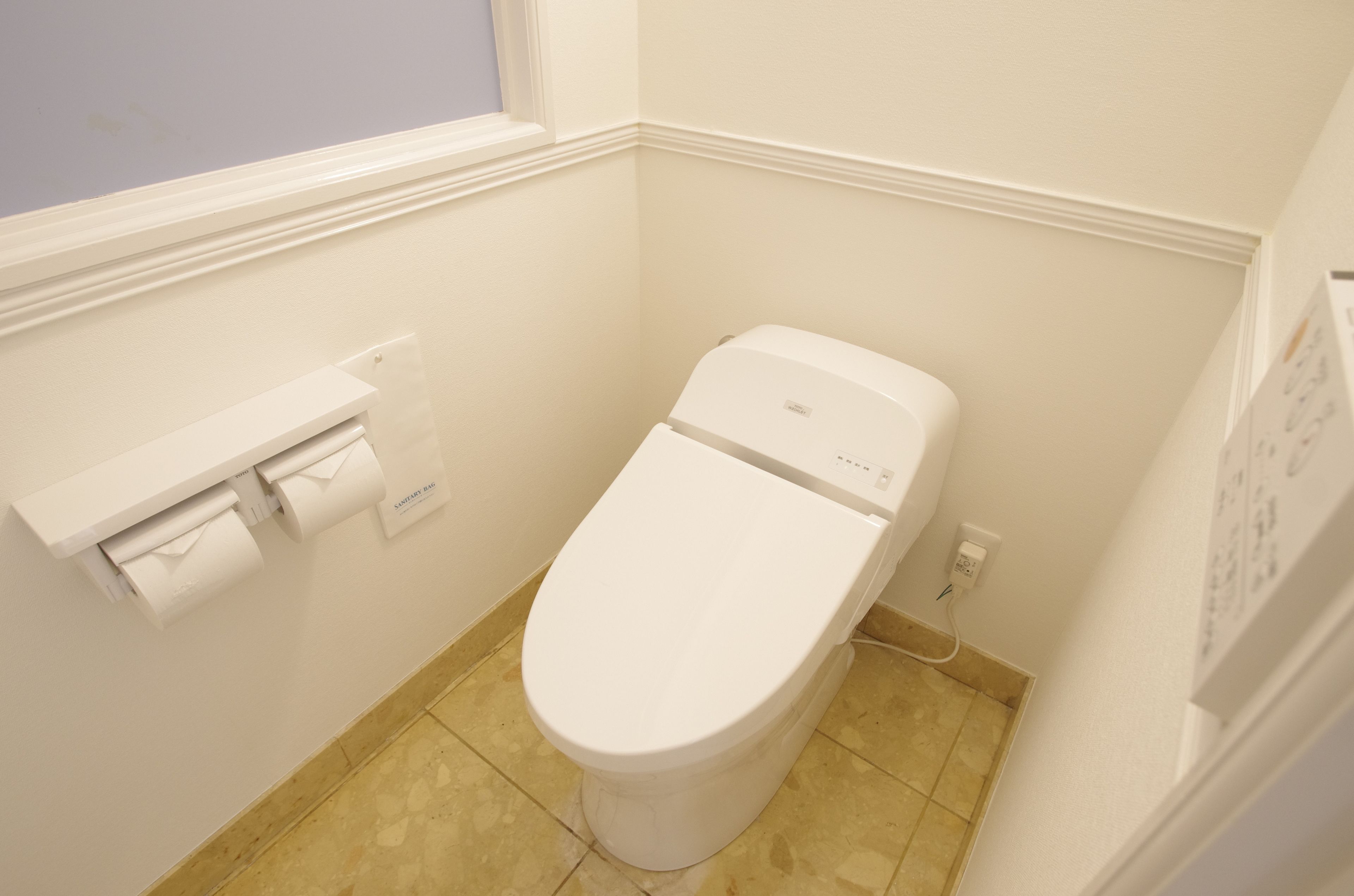 A small, white toilet in a corner bathroom with a window above it and a wall-mounted toilet paper holder to the left.
