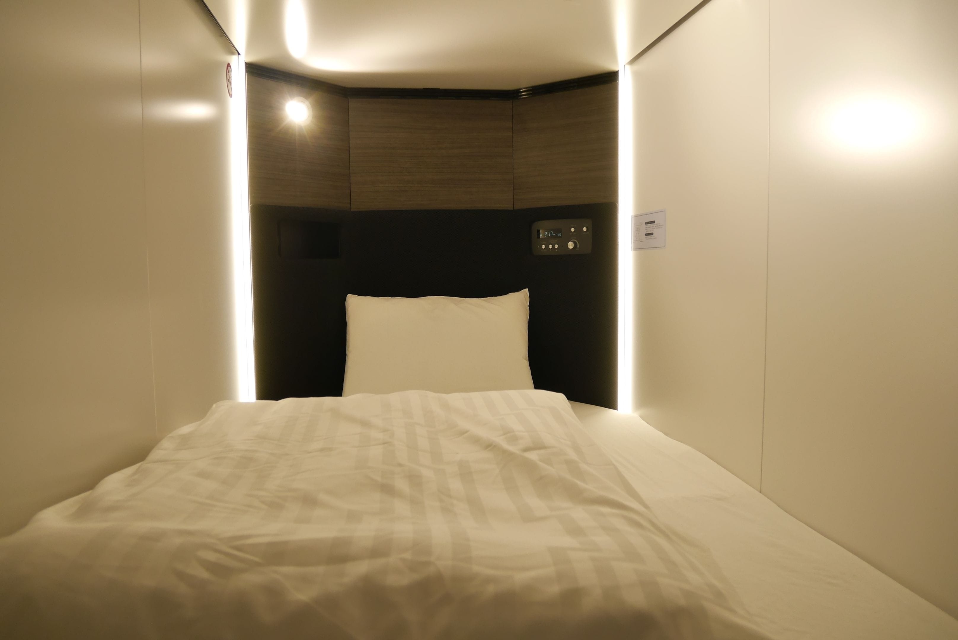Um quarto pequeno e com pouca luz, com uma cama de solteiro, um painel de controle e uma luz fixada na parede.