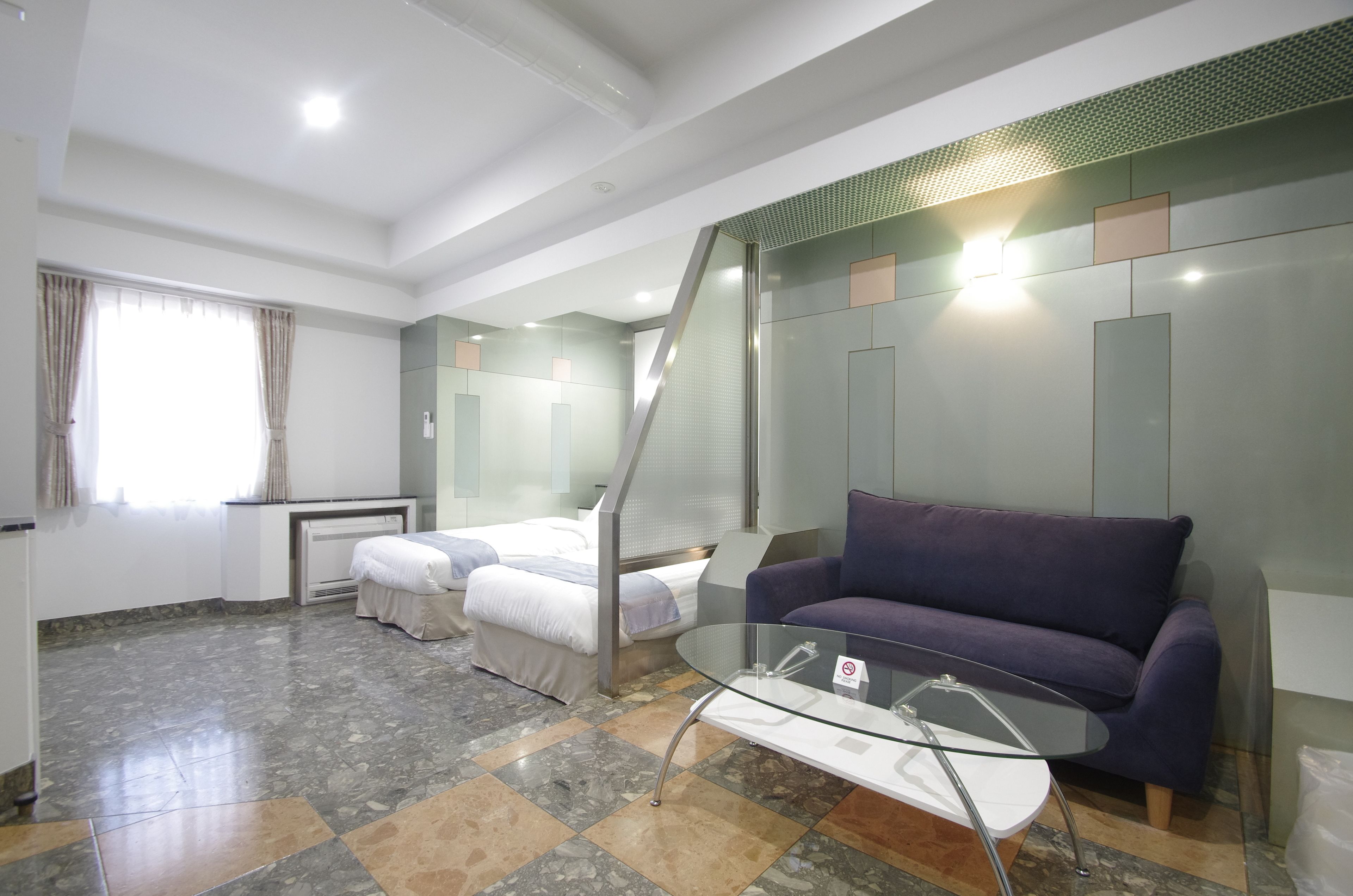 A modern hotel room with a glass coffee table, a purple sofa, a bed with white linens, and a staircase with glass railings.