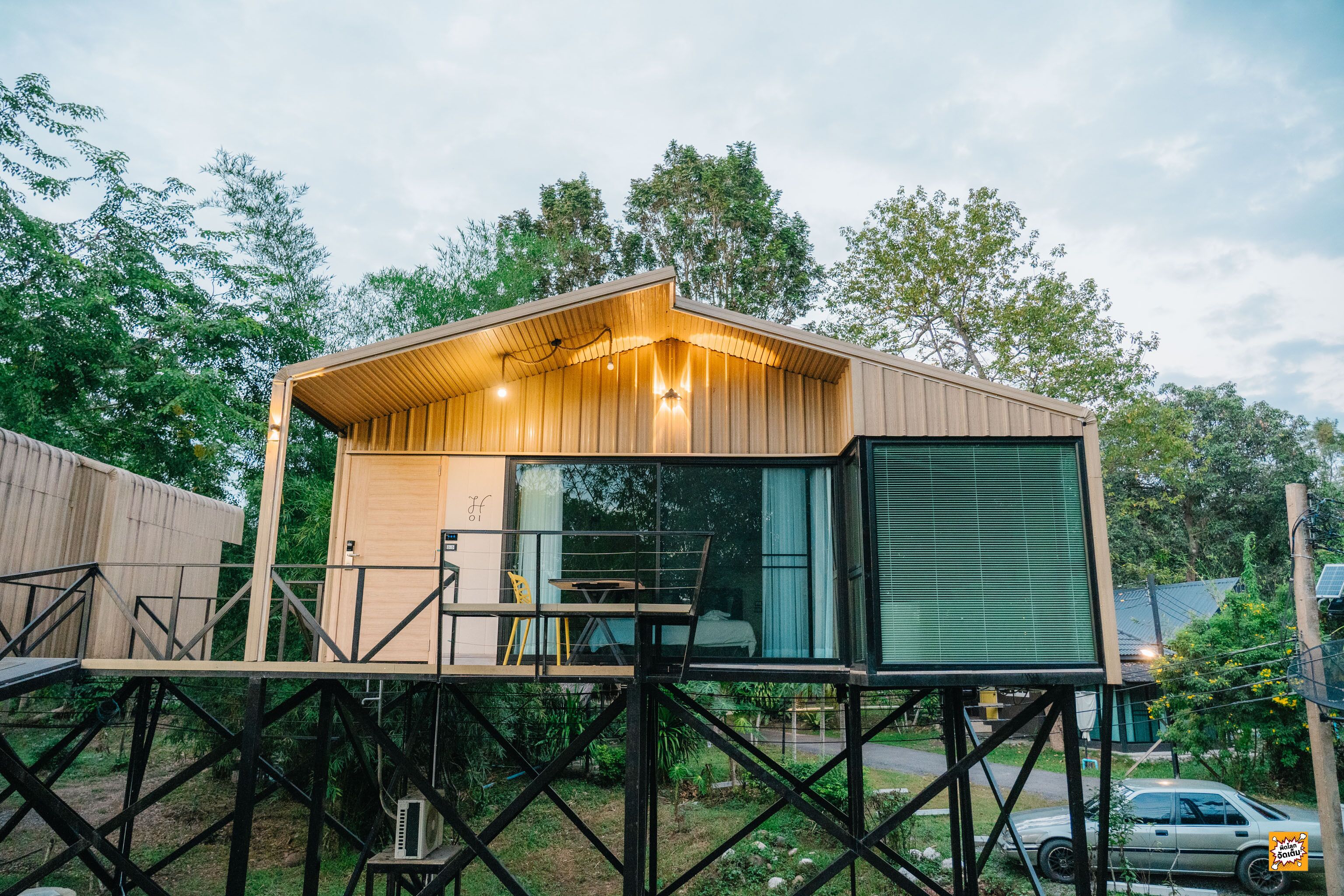 Una cabaña moderna de madera con balcón, ventanas amplias y una pequeña mesa y sillón.