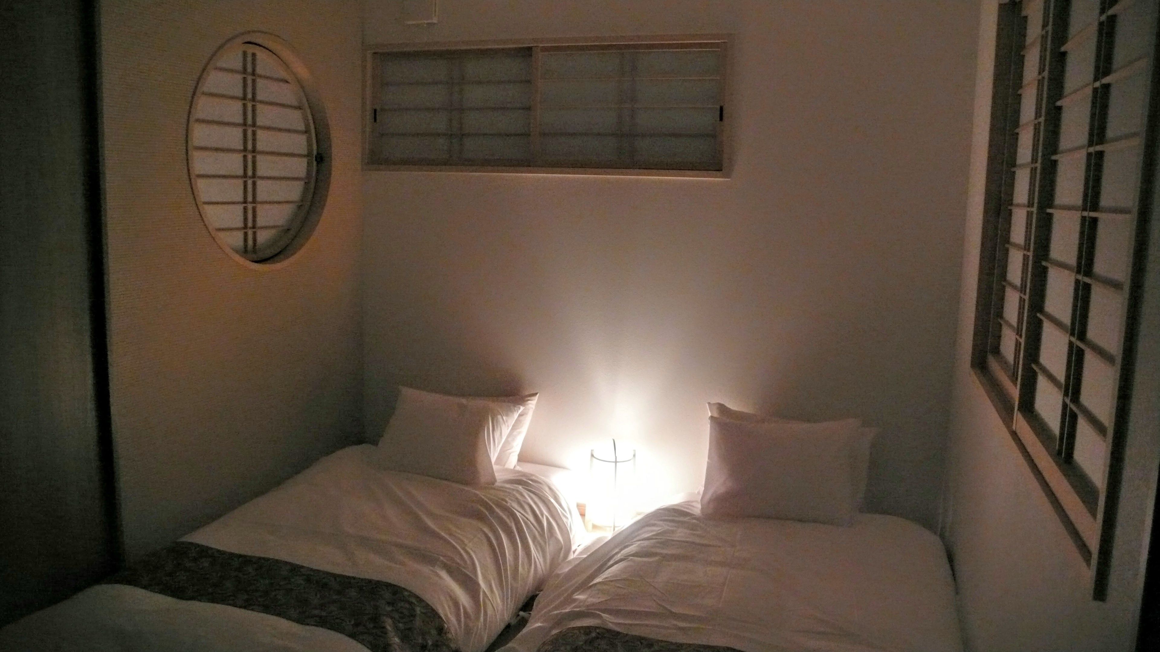 Una cama individual con sábanas blancas y dos almohadas, un espejo redondo a la izquierda y una ventana con persianas a la derecha.
