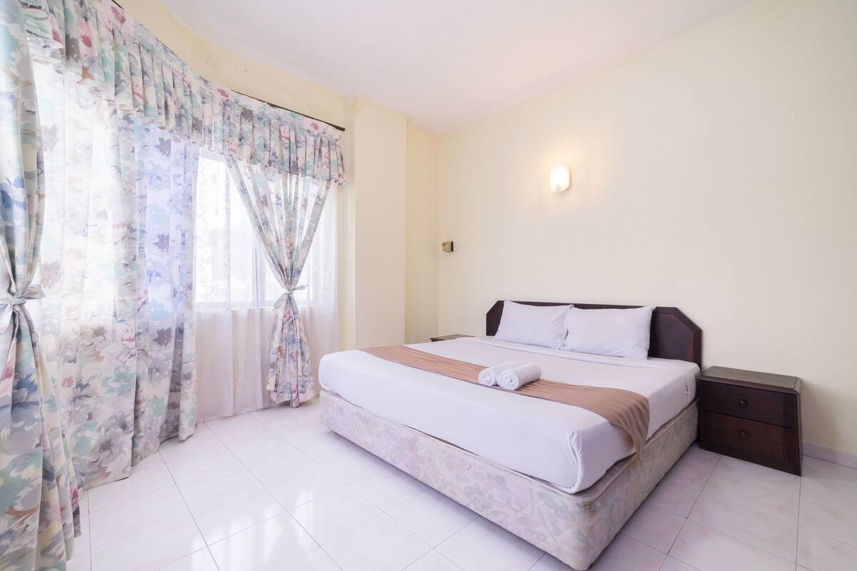 A neatly made bed with a wooden headboard, a nightstand with drawers, and a window with floral curtains.