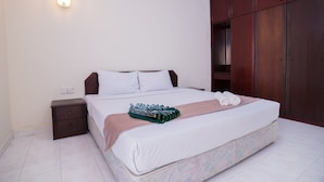 A neatly made bed with white linens, a green and pink striped blanket, and rolled towels. There is a small wooden nightstand with a drawer and a wall-mounted light fixture. -  Istana Residences Langkawi (Langkawi)
