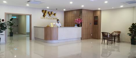 A hotel reception area with a wooden reception desk, a receptionist, and a sign for the Lipis Plaza Hotel.