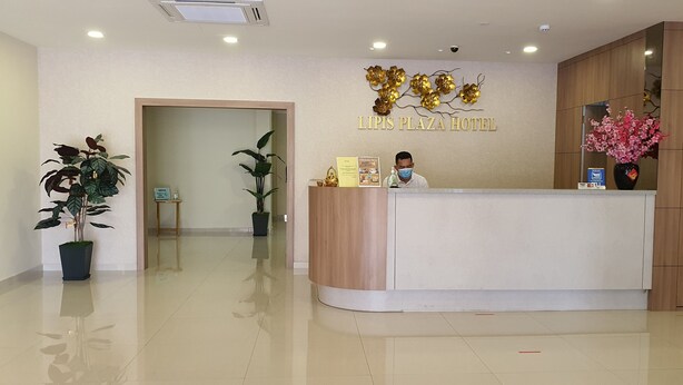 A hotel reception desk with a receptionist, a sign reading 'Lips Plaza Hotel,' and a potted plant. - Lipis Plaza Hotel (Kuala Lipis)