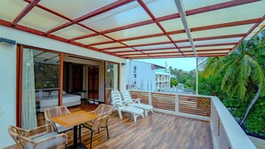 A covered outdoor seating area with a table and chairs, a wooden floor, and a view of a building and palm trees. - Kinan Retreat (Fulidhoo)