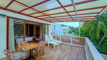 Un espacio de estar al aire libre techado con mesa y sillas, piso de madera y vista a un edificio y palmeras.