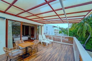Un espacio de estar al aire libre techado con mesa y sillas, piso de madera y vista a un edificio y palmeras.