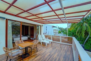 Un espacio de estar al aire libre techado con mesa y sillas, piso de madera y vista a un edificio y palmeras.