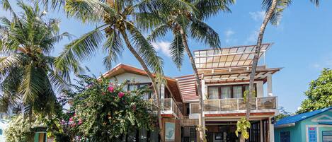 A two-story building with a balcony, surrounded by palm trees and a sandy ground.