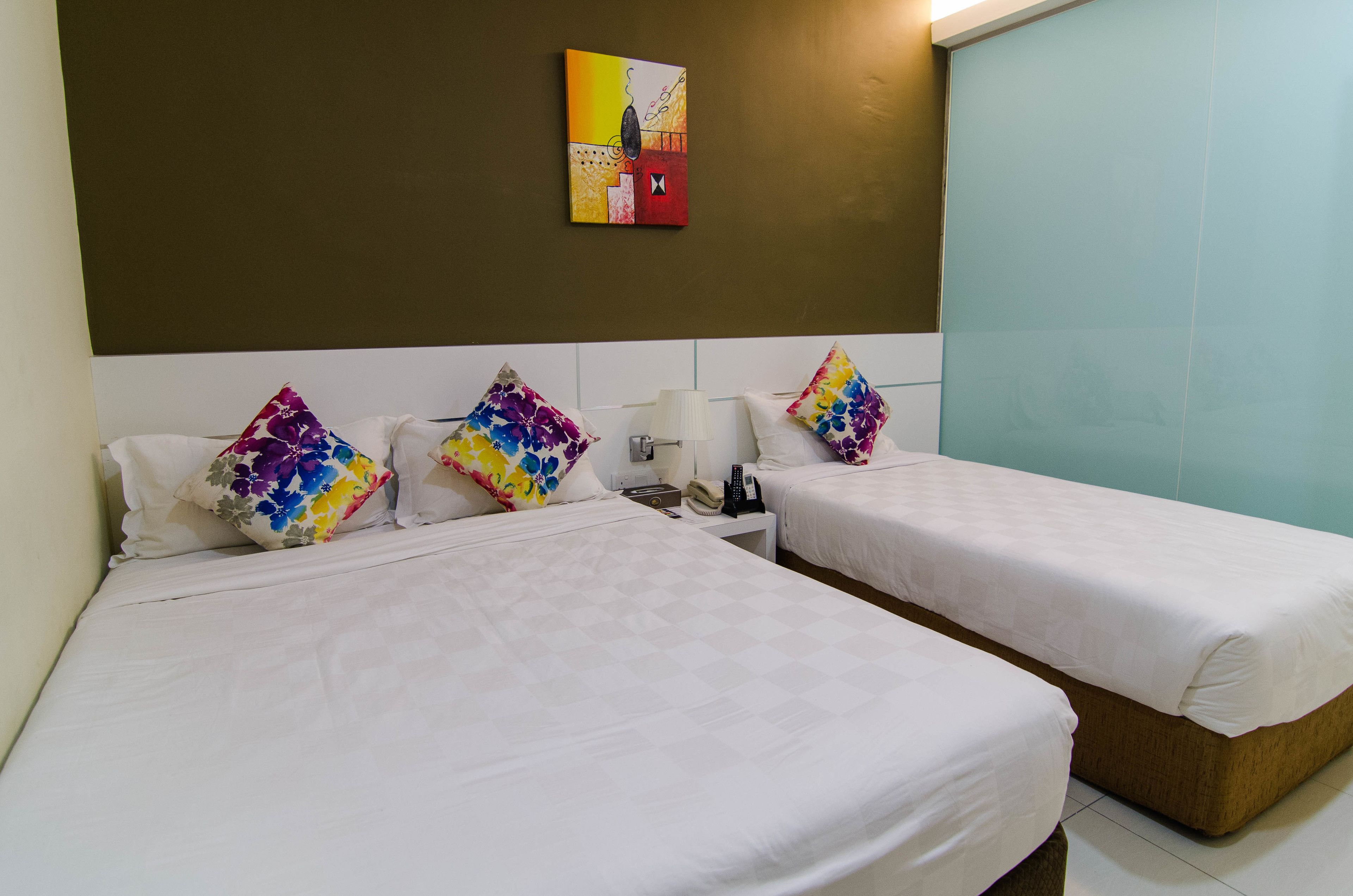 Une chambre d’hôtel avec deux lits, chacun agrémenté de draps blancs et d’oreillers aux motifs floraux colorés. On y trouve une petite table de chevet avec une lampe et un téléphone.