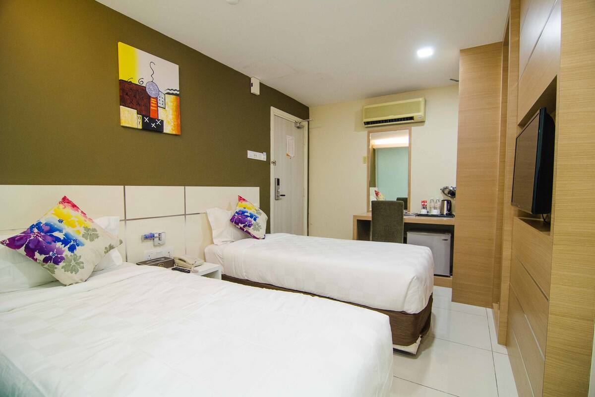 A hotel room with two beds, a desk, a chair, a television, and a small kitchenette.