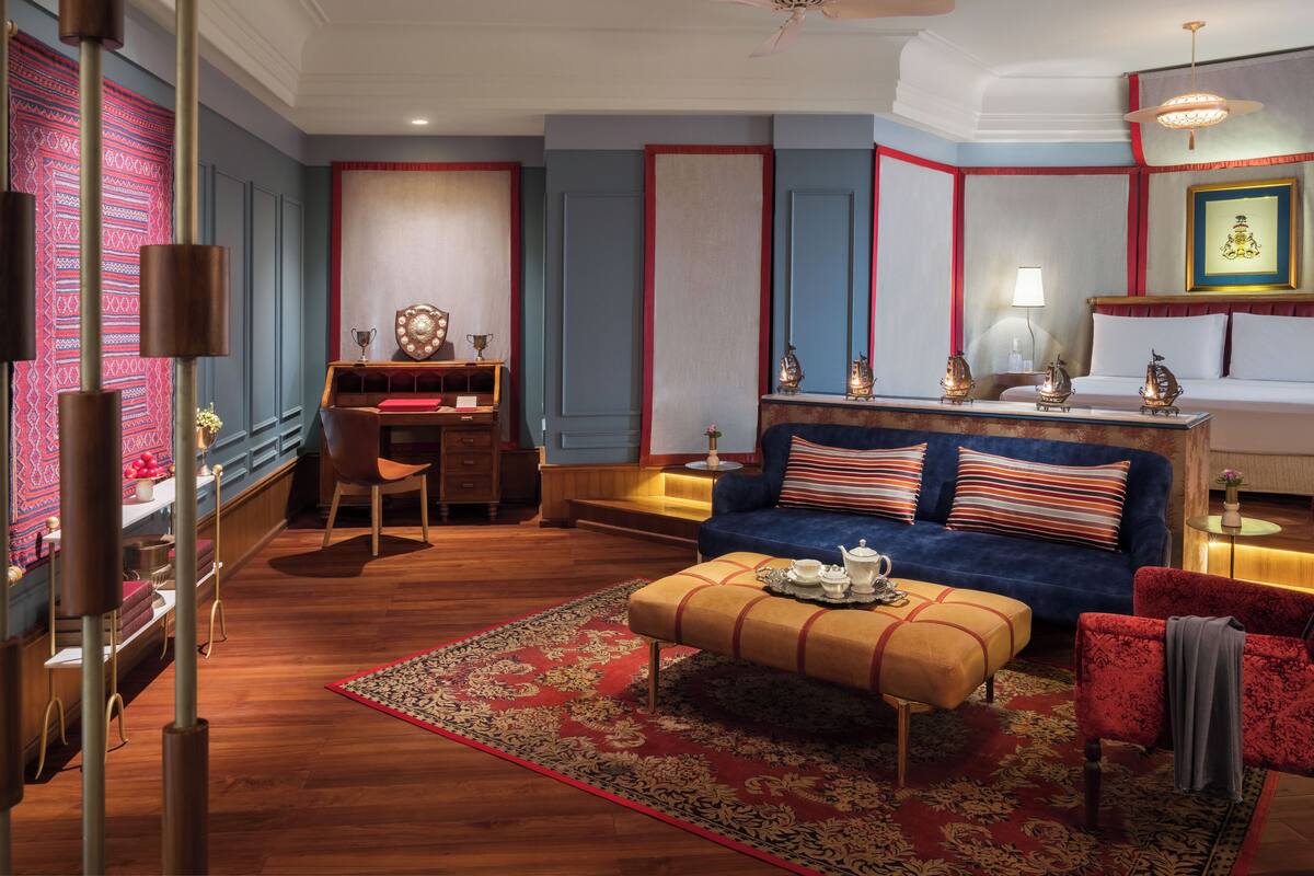 Une chambre d’hôtel avec un canapé bleu, un bureau en bois, un tapis aux motifs rouges et dorés et un ventilateur de plafond.