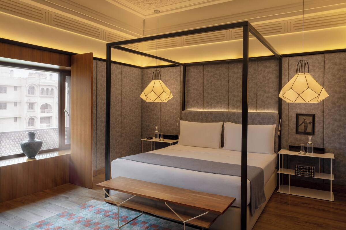 Une chambre moderne avec un lit à baldaquin, deux lampes suspendues, une table de chevet, un banc et une vue sur les bâtiments par la fenêtre.
