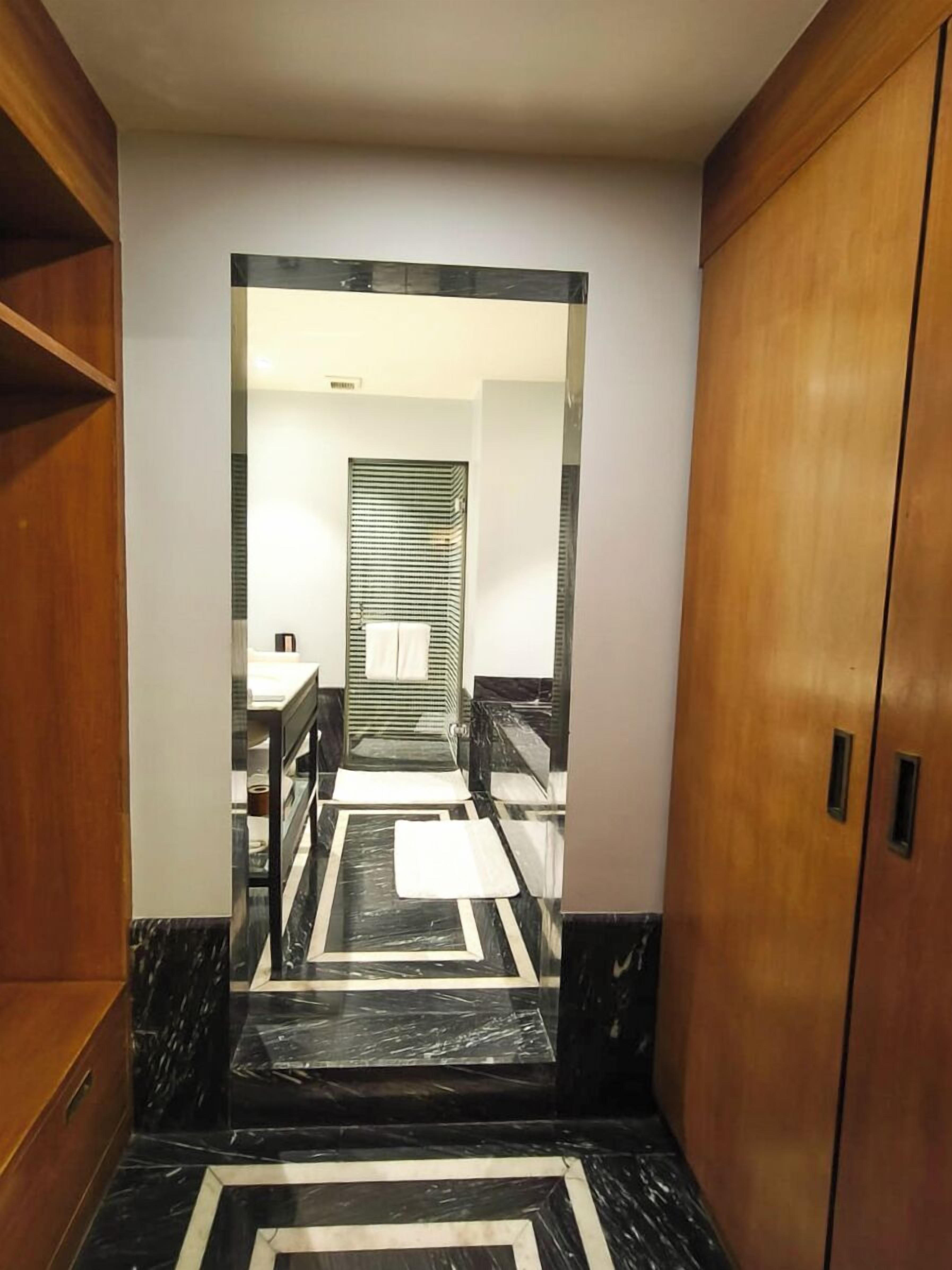 Een gang met een houten garderobe aan de rechterkant, een zwart-wit marmeren vloer, en een badkamer met een gestreept douchegordijn op de achtergrond.