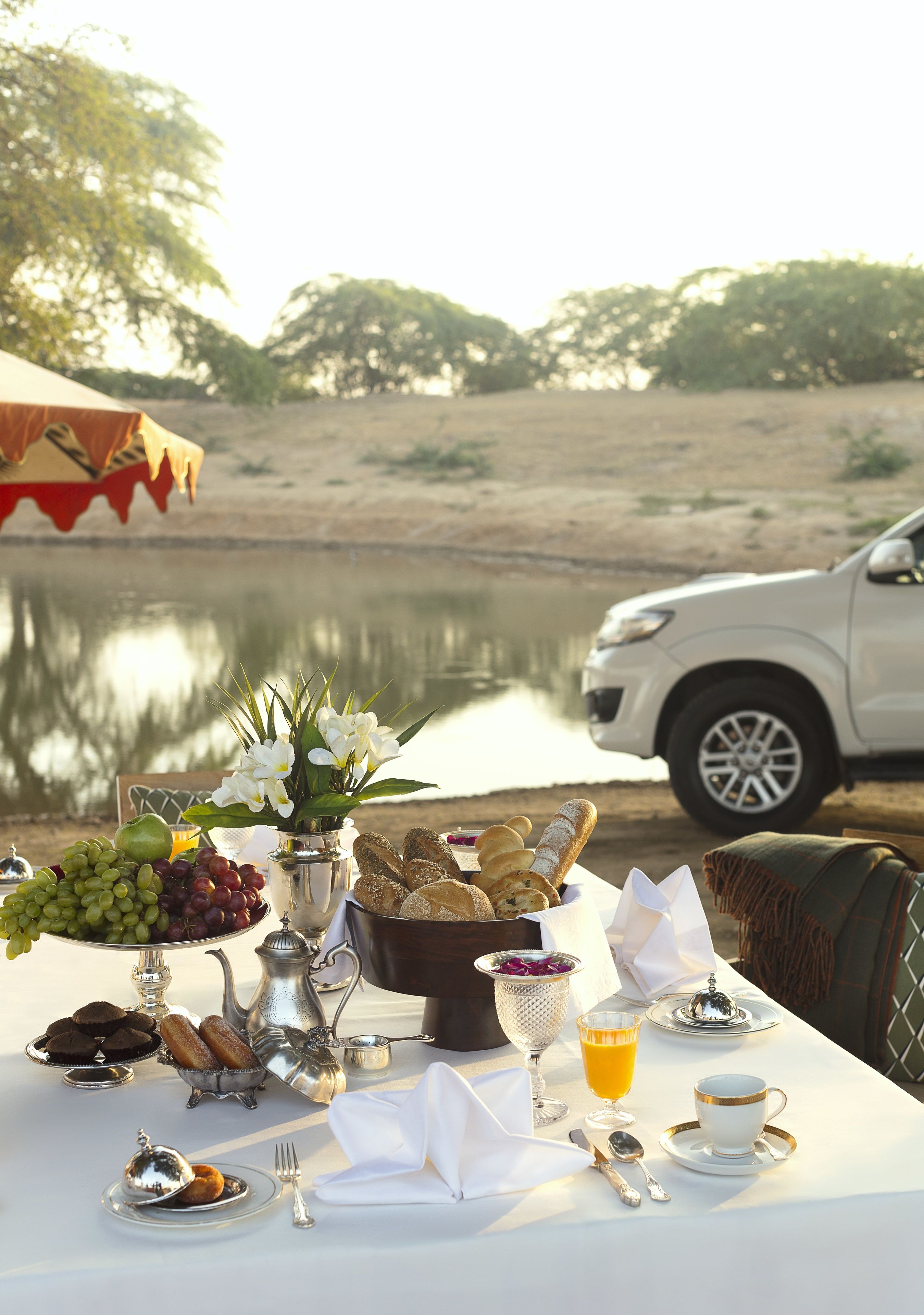 A table set with breakfast items, including fruits, bread, and a cup of coffee, by a lake with a car in the background.