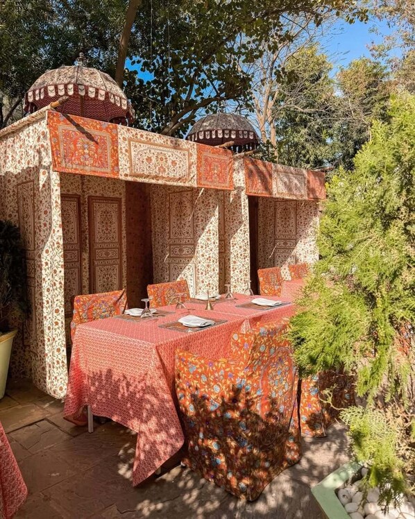 Un área de comedor al aire libre con mantel estampado, un espacio de estar cubierto con diseños intrincados y un fondo de árboles.
