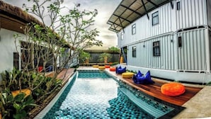 A modern outdoor pool area with a thatched-roof structure, a white building with windows, and a container-style building with a metal roof. - Us Hostel Samui (Koh Samui)