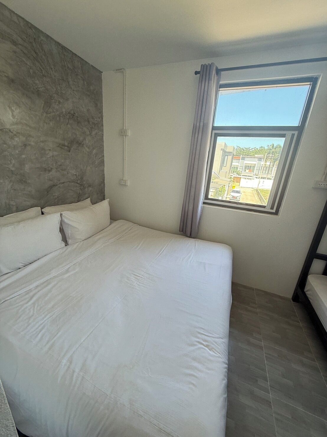 A bedroom with a large bed, a window with a view of buildings, and a grey textured wall.