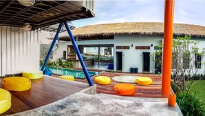 A modern building with a thatched roof, a covered outdoor area with wooden flooring and colorful bean bags, and a well-maintained lawn. - Us Hostel Samui (Koh Samui)
