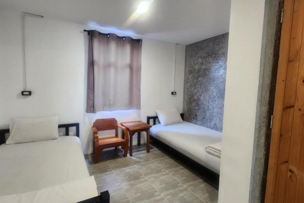 A small, well-lit room with two beds, a wooden chair, a small table, and a window with curtains.