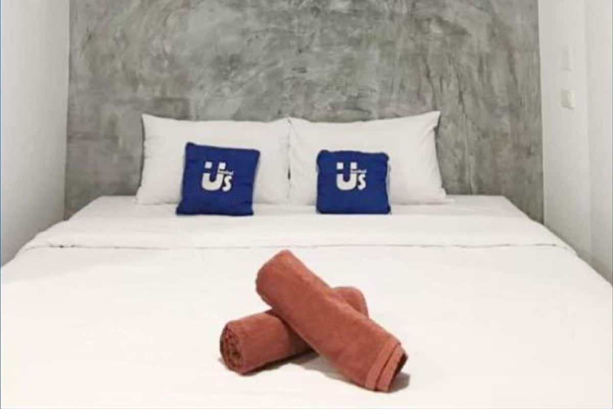 Una cama con ropa de cama blanca, dos almohadas azules con un logotipo blanco y una toalla marrón enrollada sobre la cama.