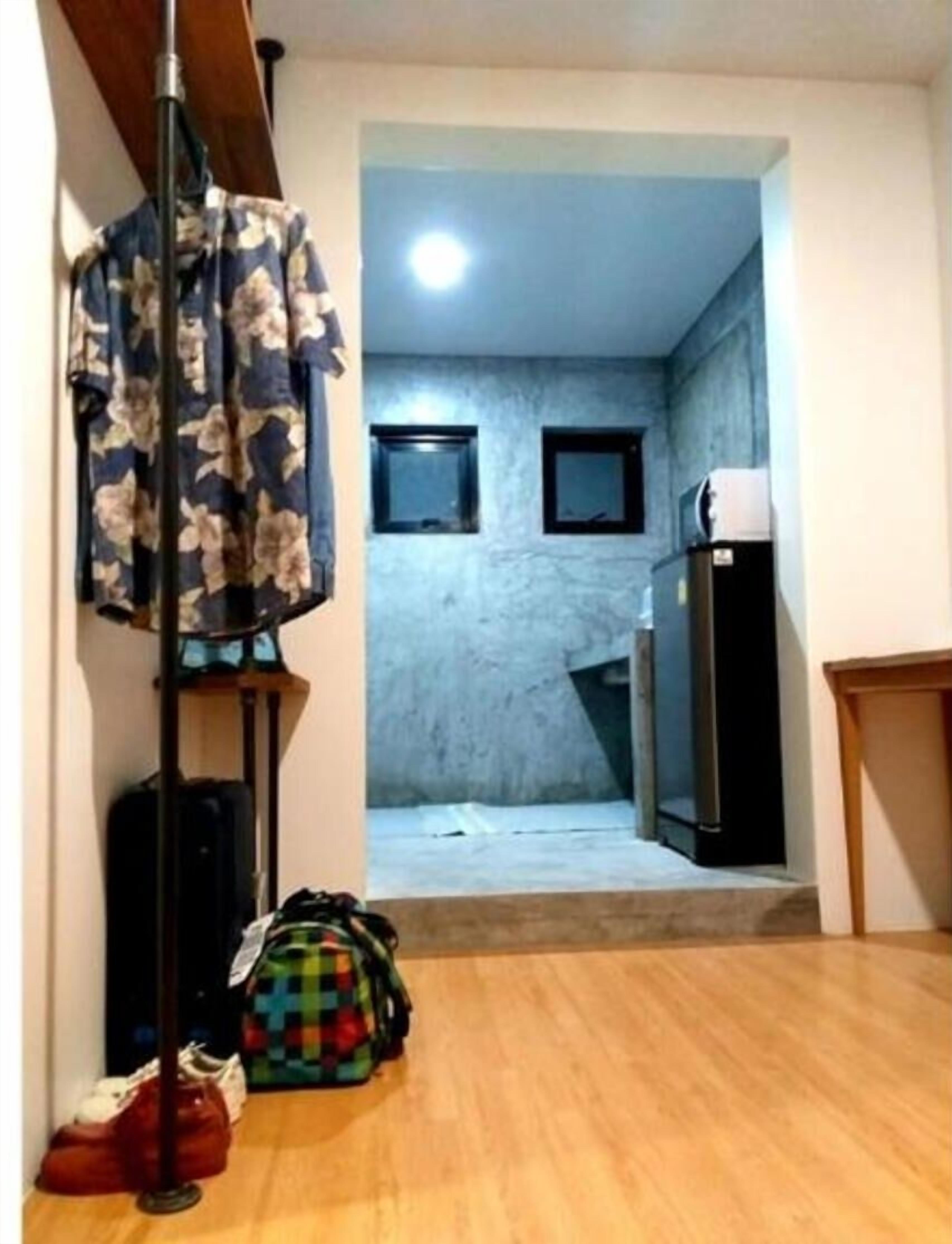 Un pasillo estrecho con suelo de madera que conduce a una zona de cocina con refrigerador y microondas. Hay una bolsa colorida en el suelo y una prenda floral colgada en un perchero.