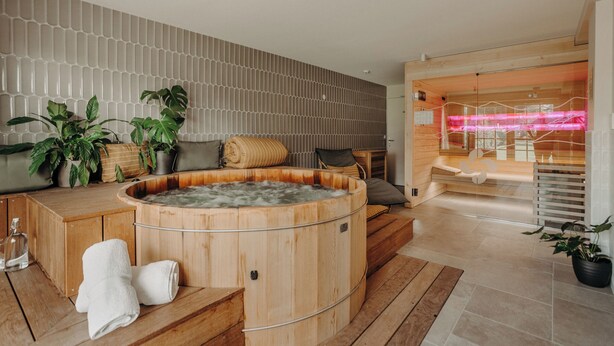 Un moderno espacio de spa con una bañera de hidromasaje de madera, suelo de madera y una zona de estar con cojines.