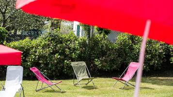 Three beach chairs and a red umbrella in a grassy area with a hedge in the background.