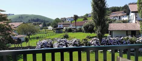 A terrace with wicker furniture, a table, and a view of a green landscape with houses and hills.