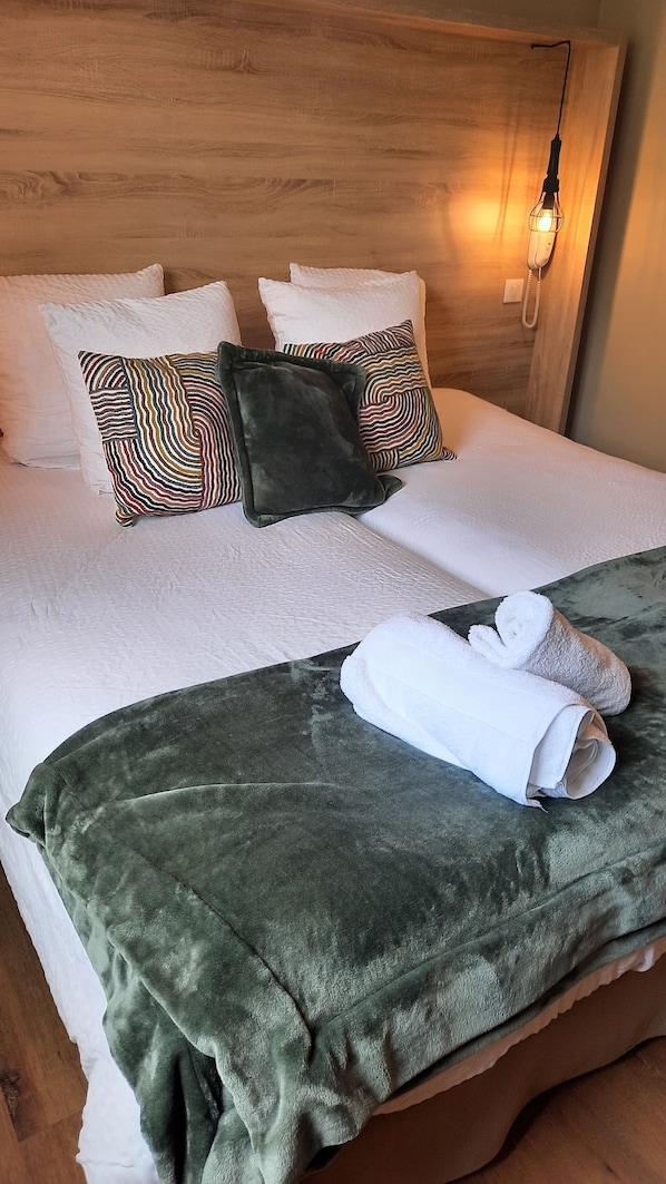 Una cama bien hecha con sábanas blancas, un plaid verde y toallas enrolladas al lado.