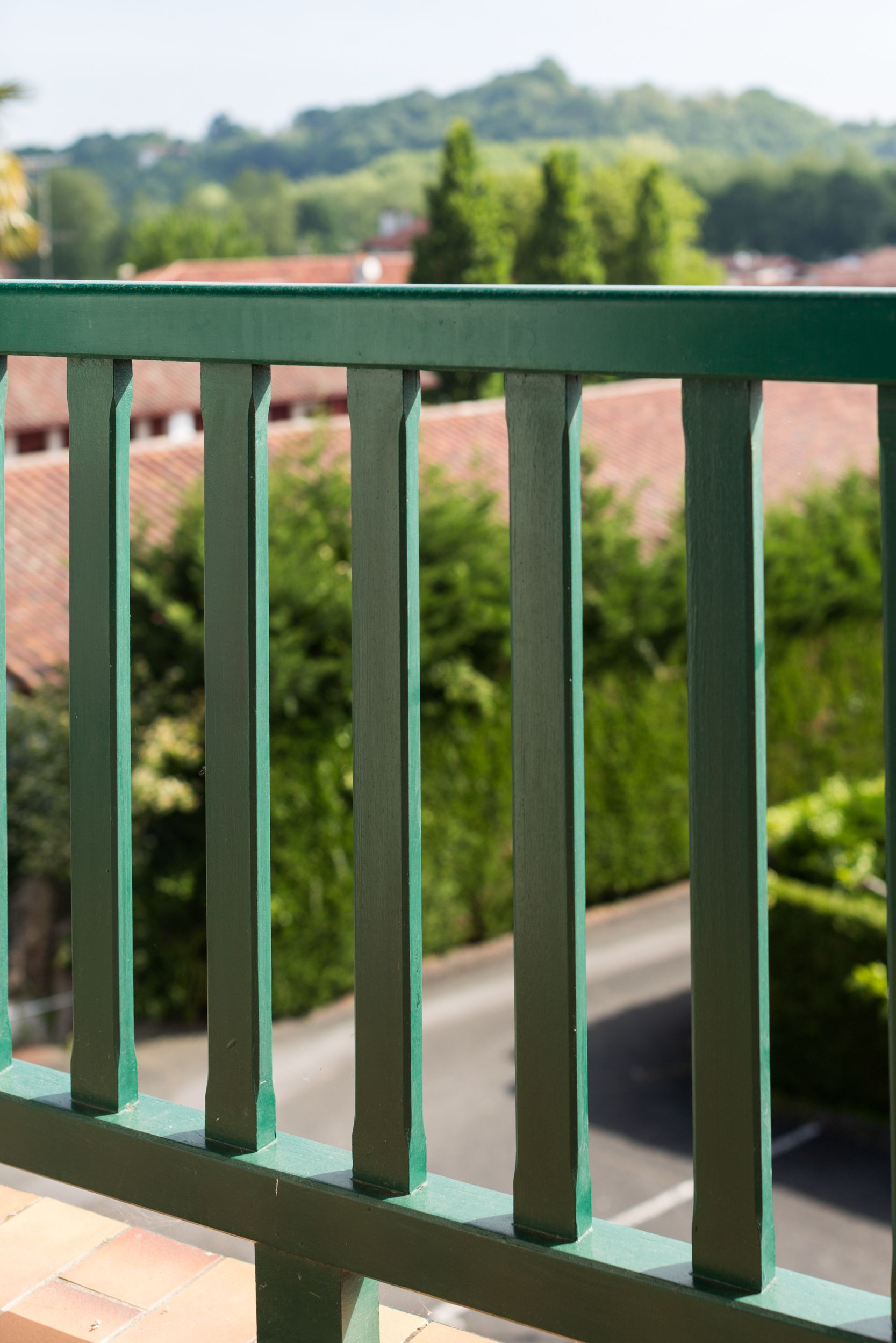 A green metal railing with a view of a road and greenery in the background.