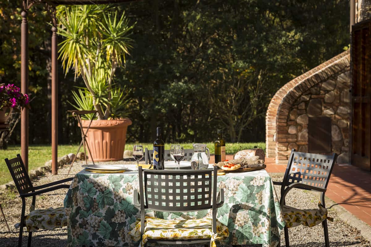 Un espace extérieur aménagé pour prendre les repas, avec une table dressée pour deux personnes, entourée de chaises, d’une plante en pot et d’un mur en pierre.