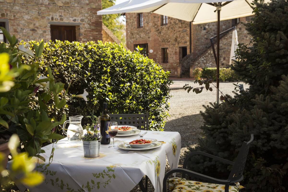 Une table à manger installée à l’extérieur, dressée avec des assiettes remplies de nourriture, une bouteille de vin et une carafe, le tout entouré de verdure et avec un bâtiment en pierre en arrière-plan.
