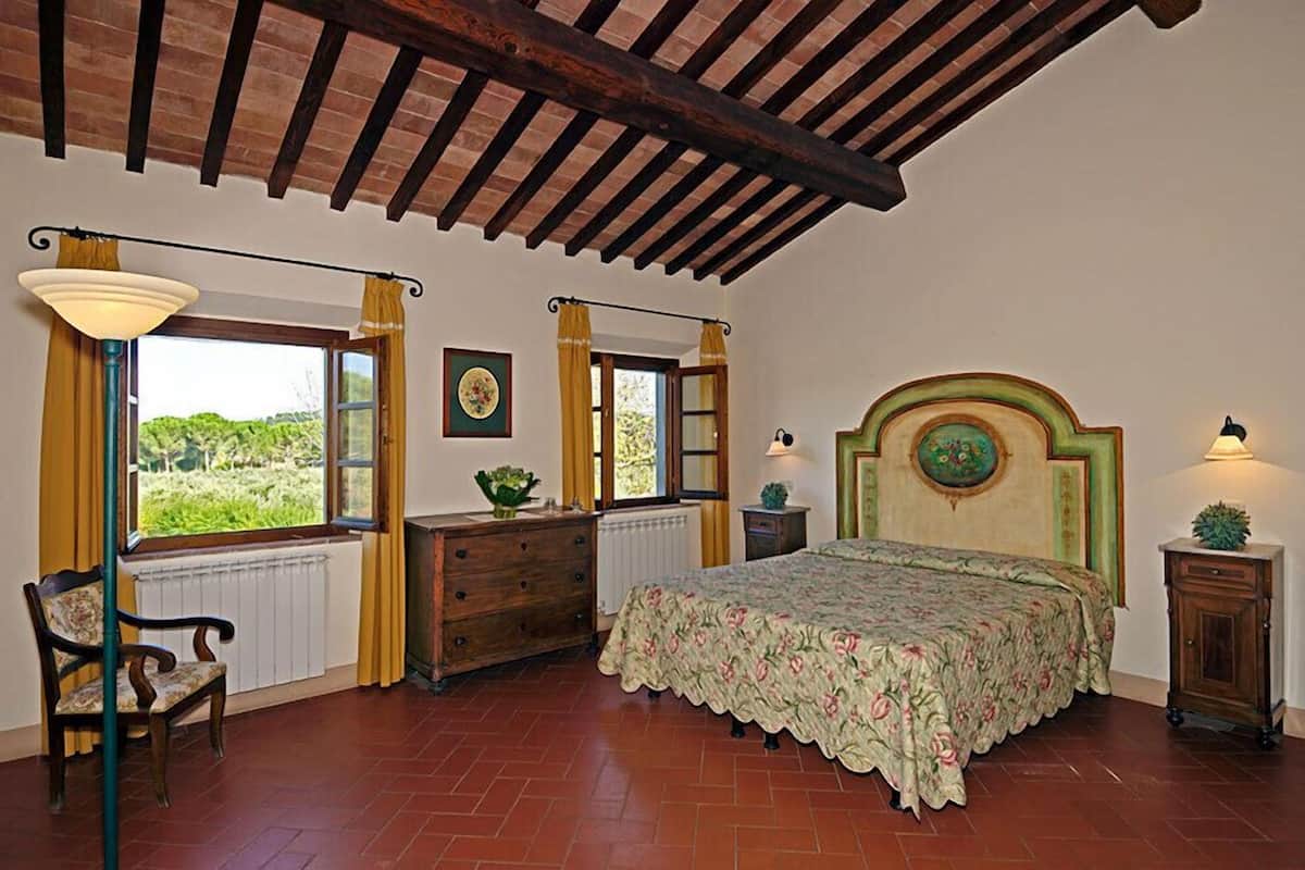Une chambre avec un plafond en bois, un lit recouvert d’une couvre-lit à motifs floraux, une chaise, une commode et deux fenêtres avec des rideaux.