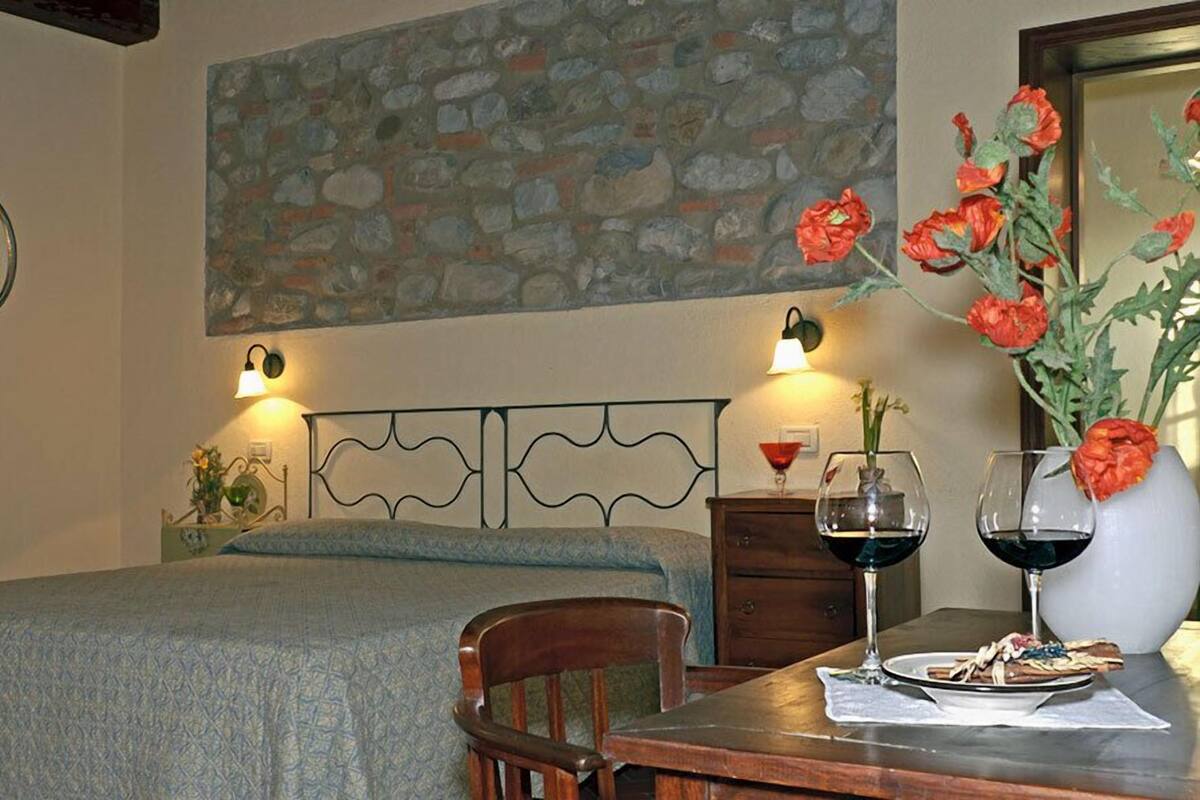 Une chambre à coucher avec un lit, une chaise en bois, une table avec une assiette de nourriture et des verres à vin, un vase avec des fleurs et une peinture murale représentant un mur de pierres.